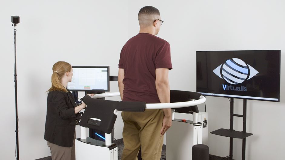 The patient is facing a monitor showing the Virtualis logo. A clinician is stood next to the patient looking at a computer screen with the test software.