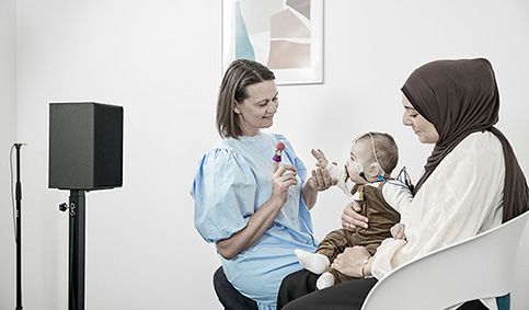 Clinician engaging infant with silent finger puppet while the aided cortical test is running. The infant is sat on their parent's lap, has electrodes placed on their head, and is facing toward the clinician and a speaker.